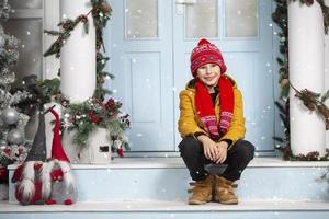 un hermoso pequeño chico en brillante ropa se sienta en el porche de un casa decorado con Navidad decoraciones foto