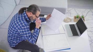 bello sorridente mezzo anziano uomo seduta su un' divano Lavorando su un' creativo progetto idea. mezzo anziano uomo utilizzando il computer portatile computer a casa Lavorando a distanza. video