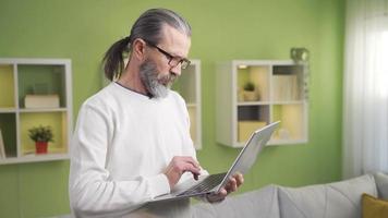 mogna man arbetssätt på Hem skriver på tangentbord på bärbar dator medan stående. mogna man använder sig av bärbar dator stående i levande rum på Hem arbetssätt, skriver på tangentbord. video