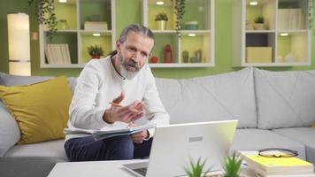 Mature man working from home discussing work on laptop video chat. Mature man talking on laptop at video conference meeting while taking notes in notebook in living room at home.