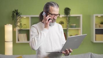 Mature man talks on the phone using a laptop at home. Business speech, meeting, brainstorming. Mature man using laptop standing in living room conducts business conversation by phone. video