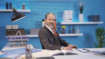 Mature businessman posing for camera with confidence and professionalism in his office. Successful and happy businessman sitting at his desk in his office smiling at the camera. video