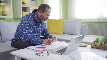 medio Envejecido Lanza libre hombre trabajando en computadora a hogar. grave y enfocado maduro hombre desarrollando nuevo proyecto mientras trabajando en ordenador portátil a hogar. video