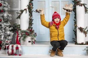 Handsome little boy in christmas. Funny child holds old skates and sits on the porch of a house decorated with Christmas tree decorations photo