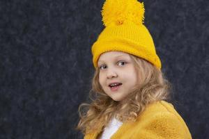 A beautiful little blonde in a knitted yellow hat looks at the camera and smiles. Happy beautiful child about five years old. photo