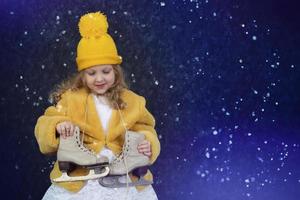 Happy little girl in a yellow fur coat and a knitted hat with skates on a blue background sprinkled with snow. photo