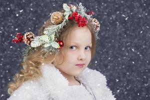 hermosa pequeño rubia niña con un Navidad guirnalda en su cabeza en un gris antecedentes con que cae nieve. Navidad niño. foto