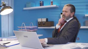 riflessivo uomo d'affari utilizzando il computer portatile e è insoddisfatto con opera. immotivato maturo uomo d'affari seduta a il suo scrivania nel il ufficio non soddisfatto con il suo opera. video