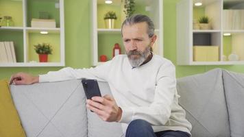 simpático velho homem sentado em sofá às casa e mensagens de texto em Smartphone. feliz velho homem mensagens de texto em a telefone às lar. video