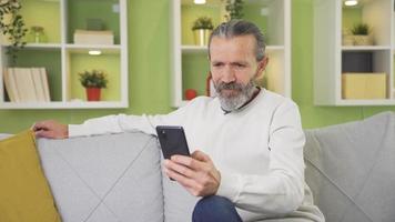 comprensivo vecchio uomo seduta su divano a casa e sms su smartphone. contento vecchio uomo sms su il Telefono a casa. video
