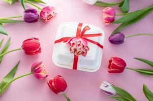 Colorful red double tulips lie around a white dessert gift box with a red ribbon. View from above photo