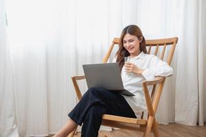 retrato de un hermosa joven mujer en un blanco camisa participación un vaso de Leche y un ordenador portátil a tipo su propio historia y sentado en un de madera silla dentro el casa, el concepto de descanso foto