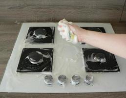 Cleaning dirty gas stove, foam from household chemicals. Hands hold a washing sponge in the foam. White glass hob surface. The concept of household chores, house cleaning. photo