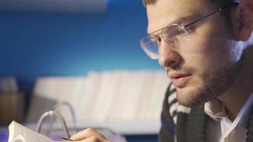 de cerca de joven hombre en lentes leyendo un libro, haciendo investigación, mirando para información. hombre haciendo investigación a hogar utilizando libros y cuadernos, culto joven hombre coleccionar información datos. video