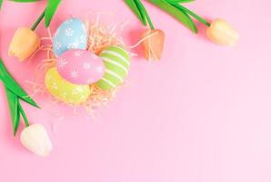 Happy Easter holiday greeting card concept. Colorful Easter Eggs and spring flowers on pastel pink background. Flat lay, top view, copy space. photo