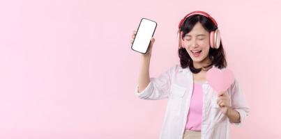 Happy cheerful smiling asian woman with hand holding heart and earphones showing blank screen mobile phone or new smartphone music application advertisement mockup isolated on pink studio background. photo