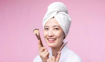 Portrait happy young asian woman with natural make up face holding cosmetic skin powder blusher isolated on pink background. Female apply skincare brush treatment. beauty product, cosmetology concept. photo