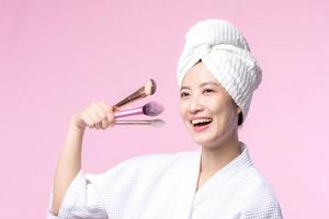 Portrait happy young asian woman with natural make up face holding cosmetic skin powder blusher isolated on pink background. Female apply skincare brush treatment. beauty product, cosmetology concept. photo