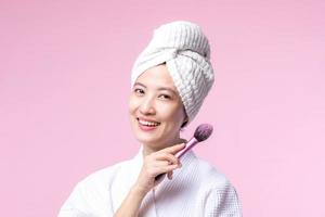 Portrait happy young asian woman with natural make up face holding cosmetic skin powder blusher isolated on pink background. Female apply skincare brush treatment. beauty product, cosmetology concept. photo