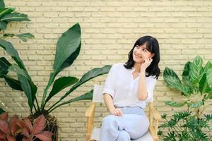 Portrait beautiful young asian woman happiness sitting on yellow cement texture grunge wall brick and green plant background, Happy female is smiling on concrete, wellness people concept. photo