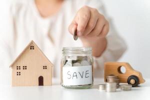 Close up of woman hand putting coin into glass jar for saving money, stack of coins, toy house and car on table, saving money and financial concept photo