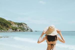 backside young woman skin tan in sunhat and bikini standing with her arms raised to her head enjoying on the beach vacation travel. and enjoy life at sea looking view of beach ocean on hot summer day. photo