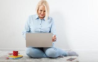 Happy middle aged woman sitting relaxed on the floor using laptop for entertainment. The concept of leisure and work with a cup of coffee at home.