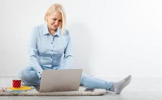 Happy middle aged woman sitting relaxed on the floor using laptop for entertainment. The concept of leisure and work with a cup of coffee at home. photo