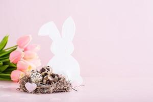 Happy easter quail eggs in a nest, white rabbit and tulips on a pink background. photo