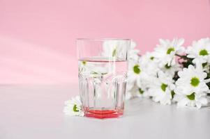 un vaso vaso con agua soportes en un rosado antecedentes con blanco crisantemo flores en el Mañana foto