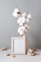 Eco friendly home decor cotton branch in a vase, photo frame and stones on the table on a gray background. Pastel shades. Vertical view