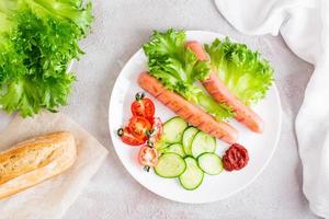 Ready-to-eat fried sausages and fresh vegetables on a plate on the table. Hearty homemade snack. Vertical view. Top view photo