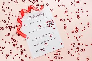 Valentine's Day. Small red hearts highlight the date February 14 on a calendar sheet and a red ribbon on a pink heart-strewn background. Top view photo