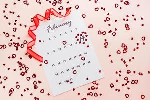 Valentine's Day. Small red hearts highlight the date February 14 on a calendar sheet and a red ribbon on a pink heart-strewn background. Top view photo
