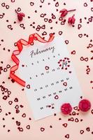 Valentine's Day. Small red hearts highlight the date February 14 on a calendar sheet, dry flowers and a red ribbon on a pink heart-strewn background. Vertical view photo