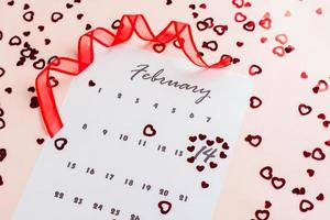 San Valentín día. pequeño rojo corazones realce el fecha febrero 14 en un calendario sábana y un rojo cinta en un rosado lleno de corazón antecedentes. de cerca foto