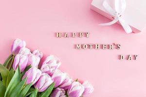 top view of a bouquet of spring tulip flowers and a festive box. text in wooden letters of Happy Mother's Day. pink background. postcard. photo