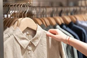 Close up of woman hand choosing thrift young and discount shirt clothes in store, searching or buying cheap cotton shirt on rack hanger at flea market. Woman choosing clothes at store. photo