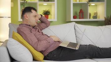 el hombre mirando a el ordenador portátil mientras acostado abajo es Deprimido por qué él ve en el pantalla. infeliz hombre acostado en vivo habitación a hogar sensación triste y suspirando mientras mirando a ordenador portátil. video