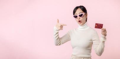 Fascinating fun joyful young woman of Asian ethnicity 20s years old with wear sunglasses wears white shirt hold in hand credit bank card isolated on plain pastel light pink background studio portrait. photo