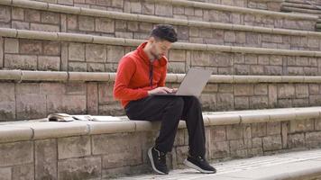 masculino aluna estudando ao ar livre parece às computador portátil e livros. uma jovem aluna é sentado em uma pedra dentro a parque e alegremente estudando com uma computador portátil. video
