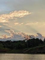 A boat is in the water and the sky is blue and the water is calm. A tree in the foreground with the sun shining through the clouds. The water is calm. photo