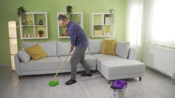 Old mature man does his own cleaning at home and gets very tired. Tiring retirement. An elderly man is mopping the floors in the living room at home for hygiene and getting tired. video