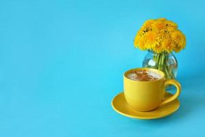 Cappuccino topped with whipped cream and cinnamon in yellow cup and bouquet of fresh dandelions in little glass vase on blue backgroung. Hello summer concept. Place for your text here. photo