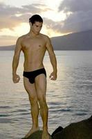 As the sun sets in the background this handsome young man at the beach in Maui, Hawaii shows off in a speedo, letting people see his muscular fit and sexy body. photo