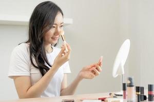 Happy beauty concept, beautiful asian young woman, girl, make up face by applying brush blush powder on her cheek, looking in mirror at home, look with natural fashion style, isolated on background. photo