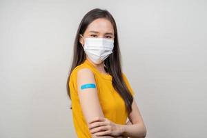 Portrait of health care asian young woman wearing face mask protect, getting vaccine of covid-19, influenza in yellow t-shirt, showing arm on blue bandage, isolated on nature background, copy space.