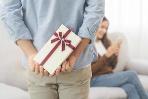 Celebrating Valentine's day anniversary, relationship two asian young couple love, boyfriend gives a gift to girlfriend by hide box at the behind, woman getting present while sitting on sofa at home. photo