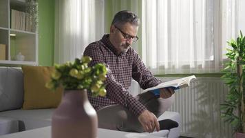 une mature homme est repos à maison, en train de lire une livre et découverte paix. âge moyen homme dans des lunettes en train de lire une livre dans le sien paisible moderne maison, profiter le sien gratuit temps. video