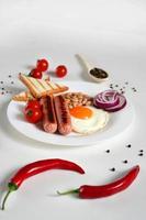 Traditional English Breakfast. Plate with Fried egg, two fried sausages, grilled bread toast, canned beans, blue onions and cherry tomatoes on a White Background. Copy space photo